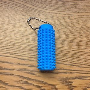 Puede incluir: Una herramienta de masaje cil&iacute;ndrica azul con una superficie texturizada de protuberancias triangulares espaciadas uniformemente. Tiene una peque&ntilde;a cadena de bolas de metal unida a la parte superior. La herramienta est&aacute; dise&ntilde;ada para el alivio muscular y mide unos 10 cm de largo.