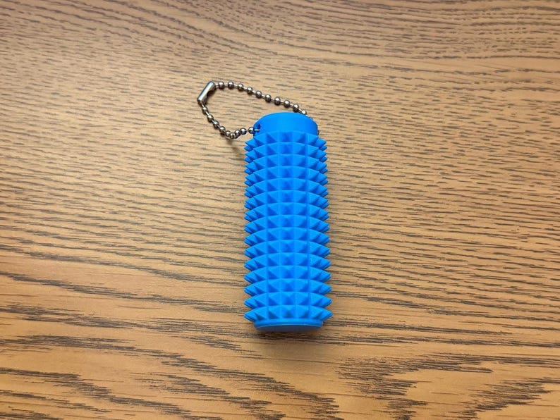 Puede incluir: Una herramienta de masaje cil&iacute;ndrica azul con una superficie texturizada de protuberancias triangulares espaciadas uniformemente. Tiene una peque&ntilde;a cadena de bolas de metal unida a la parte superior. La herramienta est&aacute; dise&ntilde;ada para el alivio muscular y mide unos 10 cm de largo.