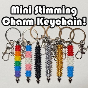 May include: Six cylindrical stimming charm keychains with spiky textures and metal hardware. The charms feature various color patterns including rainbow, pink-white-blue, solid black, solid silver, solid gold, and red-white-blue. Text reads: Mini Stimming Charm Keychain!