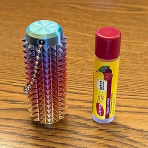 Puede incluir: Un recipiente multicolor impreso en 3D con una cadena junto a un tubo de b&aacute;lsamo labial Carmex. El recipiente tiene un dise&ntilde;o puntiagudo y degradados de azul, morado, naranja y rojo. El b&aacute;lsamo labial es amarillo con tapa roja.