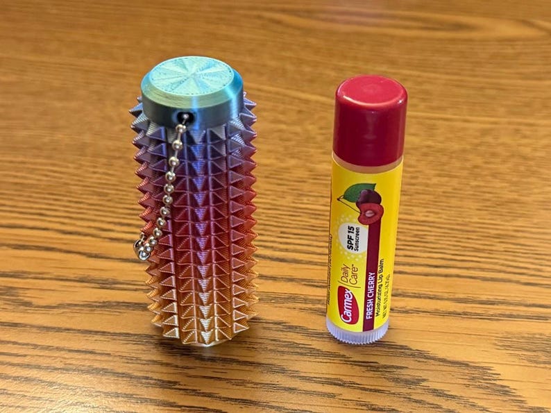 Puede incluir: Un recipiente multicolor impreso en 3D con una cadena junto a un tubo de b&aacute;lsamo labial Carmex. El recipiente tiene un dise&ntilde;o puntiagudo y degradados de azul, morado, naranja y rojo. El b&aacute;lsamo labial es amarillo con tapa roja.