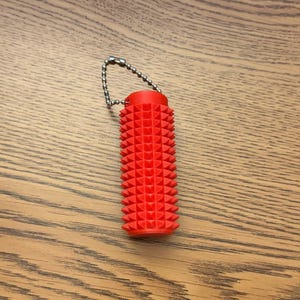 Puede incluir: Una herramienta de masaje de color rojo brillante, de forma cil&iacute;ndrica, con una superficie texturizada con p&uacute;as. Est&aacute; unida a una peque&ntilde;a cadena de metal. El art&iacute;culo est&aacute; dise&ntilde;ado para el alivio muscular y se muestra sobre una superficie de madera.