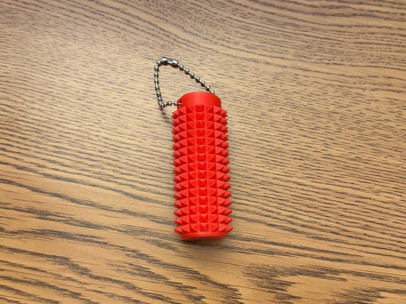 Puede incluir: Una herramienta de masaje de color rojo brillante, de forma cil&iacute;ndrica, con una superficie texturizada con p&uacute;as. Est&aacute; unida a una peque&ntilde;a cadena de metal. El art&iacute;culo est&aacute; dise&ntilde;ado para el alivio muscular y se muestra sobre una superficie de madera.