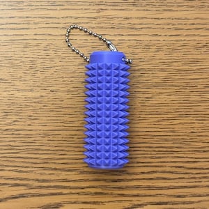 Puede incluir: Un objeto cil&iacute;ndrico texturizado de color lavanda con una cadena adjunta. El objeto est&aacute; cubierto con un patr&oacute;n de formas triangulares en relieve. La cadena est&aacute; hecha de peque&ntilde;as bolas de metal plateado. Est&aacute; sobre una superficie de madera.