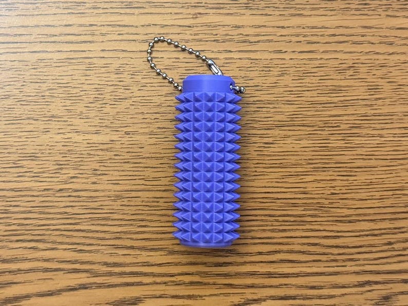 Puede incluir: Un objeto cil&iacute;ndrico texturizado de color lavanda con una cadena adjunta. El objeto est&aacute; cubierto con un patr&oacute;n de formas triangulares en relieve. La cadena est&aacute; hecha de peque&ntilde;as bolas de metal plateado. Est&aacute; sobre una superficie de madera.