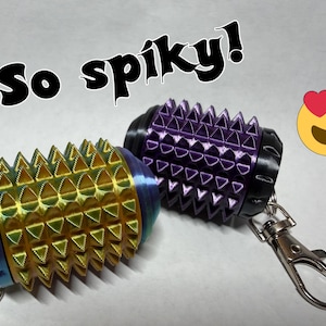 May include: Two cylindrical keychains with a spiky, textured surface. One keychain is iridescent gold and blue, while the other is purple with black end caps. Both feature metal lobster claw clasps. The text "So spiky!" and a heart-eyed emoji are visible.