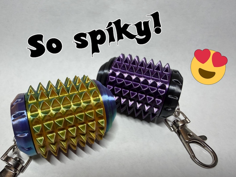 May include: Two cylindrical keychains with a spiky, textured surface. One keychain is iridescent gold and blue, while the other is purple with black end caps. Both feature metal lobster claw clasps. The text "So spiky!" and a heart-eyed emoji are visible.