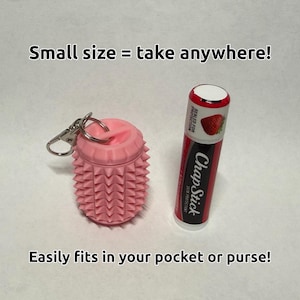 May include: A pink cylindrical silicone container with a textured spiked surface and a metal keychain ring, positioned next to a red and black ChapStick lip balm tube. The text reads: Small size = take anywhere! Easily fits in your pocket or purse!