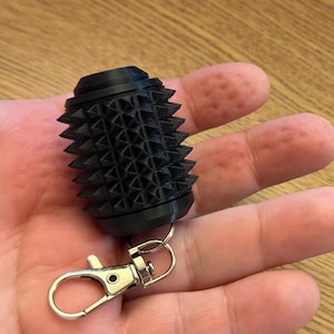 May include: A black, cylindrical keychain accessory featuring a textured surface covered in small, uniform, pyramid-shaped spikes. The object is attached to a silver-colored metal lobster clasp, which is connected by a small circular metal ring.