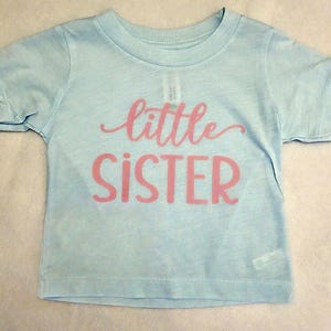 May include: A light blue short-sleeved t-shirt featuring the text little SISTER printed in pink across the chest. The shirt has a crew neckline and a simple, straight-cut design with a soft, heathered appearance in the fabric texture.