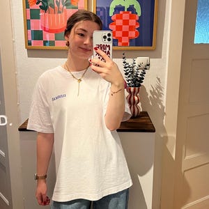 May include: A person wearing a white t-shirt with blue text and blue jeans. Two framed abstract art pieces hang on the wall behind the person. A vase with a red and white pattern sits on a wooden shelf.