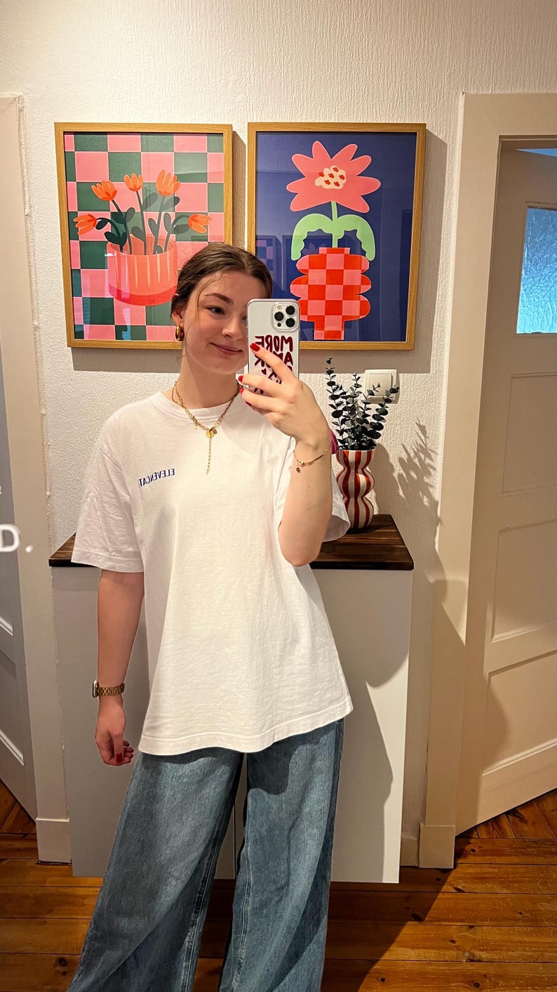 May include: A person wearing a white t-shirt with blue text and blue jeans. Two framed abstract art pieces hang on the wall behind the person. A vase with a red and white pattern sits on a wooden shelf.