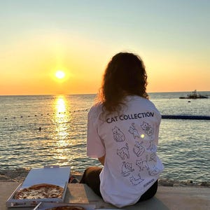 May include: A white t-shirt with a cat illustration and the text "CAT COLLECTION" is visible. Two pizza boxes are open, revealing pizzas with toppings. The scene is set at sunset, with the ocean and sky in the background.