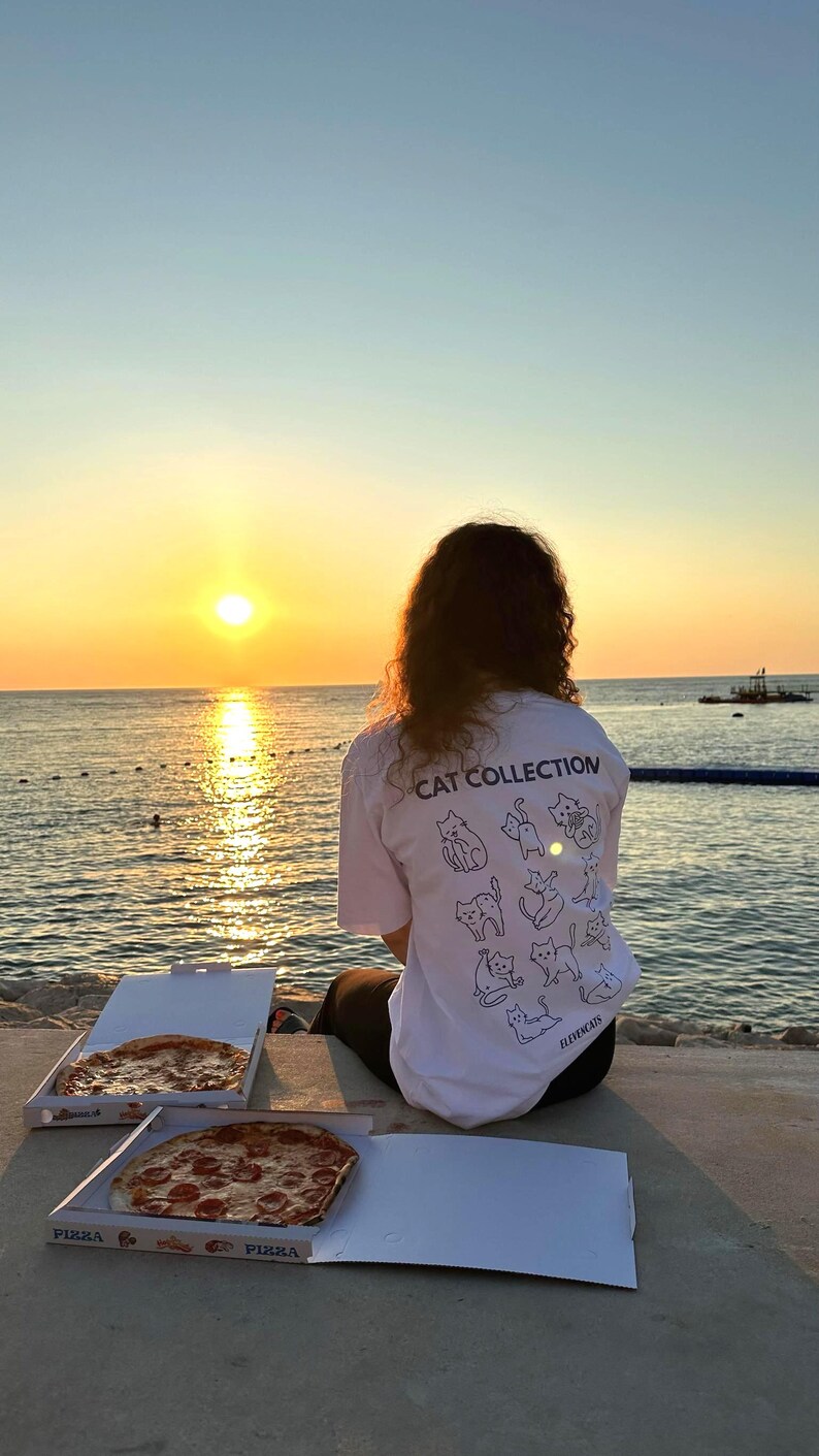 May include: A white t-shirt with a cat illustration and the text "CAT COLLECTION" is visible. Two pizza boxes are open, revealing pizzas with toppings. The scene is set at sunset, with the ocean and sky in the background.
