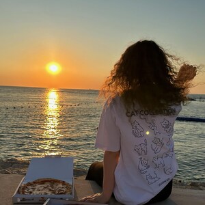 May include: A white t-shirt with a "Cat Collection" graphic and several cat illustrations on the back. Two pizza boxes with pizza are in front of a person watching the sunset over the ocean.