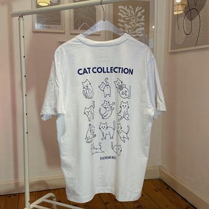 May include: White t-shirt with the text "CAT COLLECTION" and illustrations of various cat poses in blue. The shirt is hanging on a white metal clothing rack. The brand name "ELEVENCATS" is printed at the bottom.
