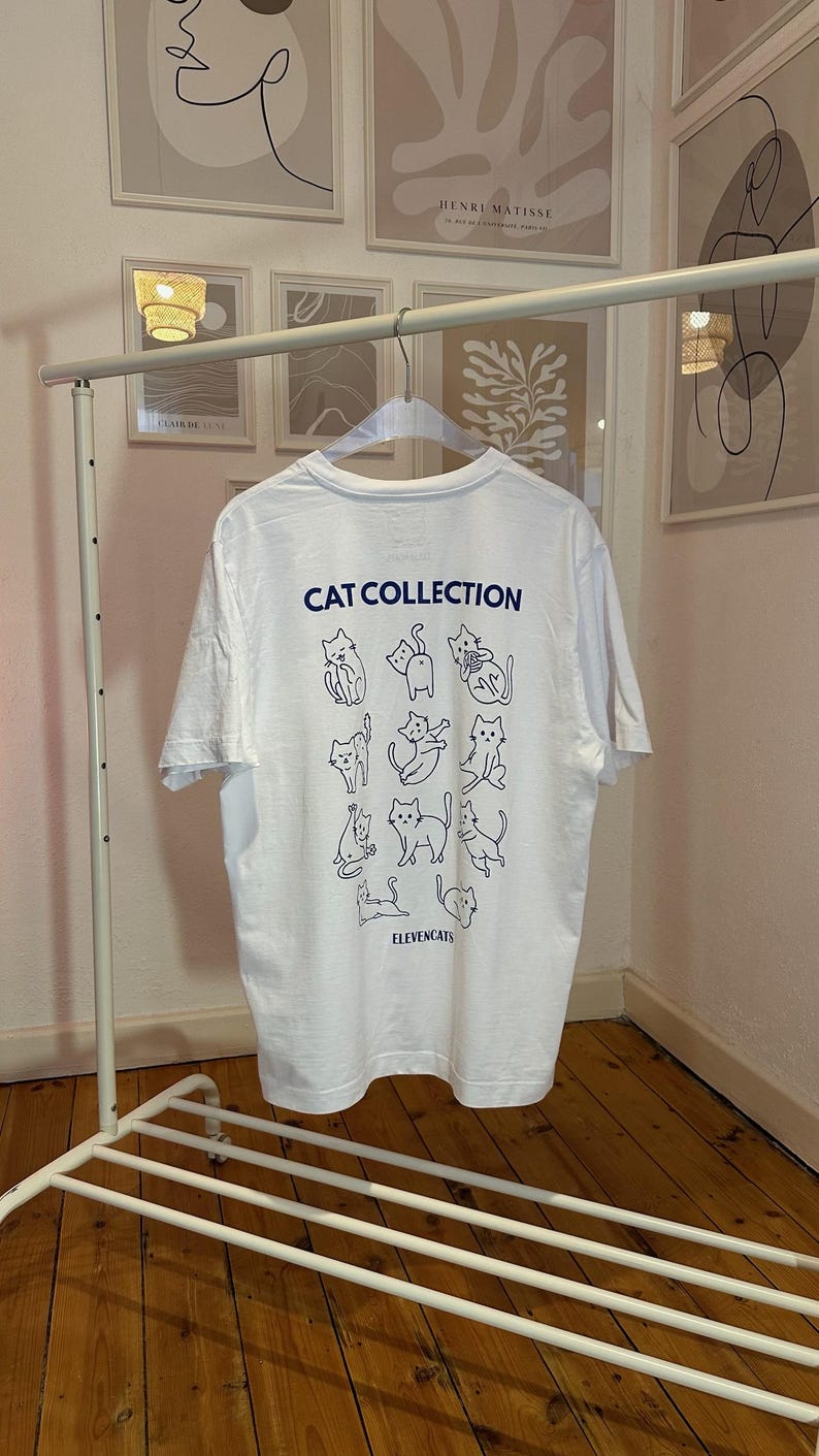 May include: White t-shirt with the text "CAT COLLECTION" and illustrations of various cat poses in blue. The shirt is hanging on a white metal clothing rack. The brand name "ELEVENCATS" is printed at the bottom.