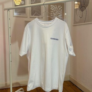 May include: A white t-shirt with the text "ELEVENCATS" in a dark blue font. The t-shirt is hanging on a white metal clothing rack. Artwork is visible in the background.
