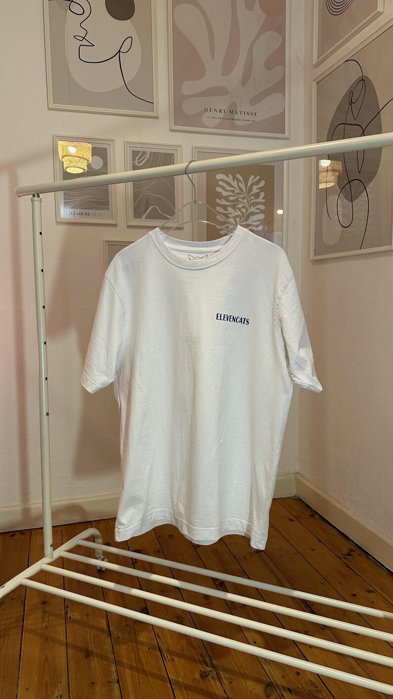 May include: A white t-shirt with the text "ELEVENCATS" in a dark blue font. The t-shirt is hanging on a white metal clothing rack. Artwork is visible in the background.