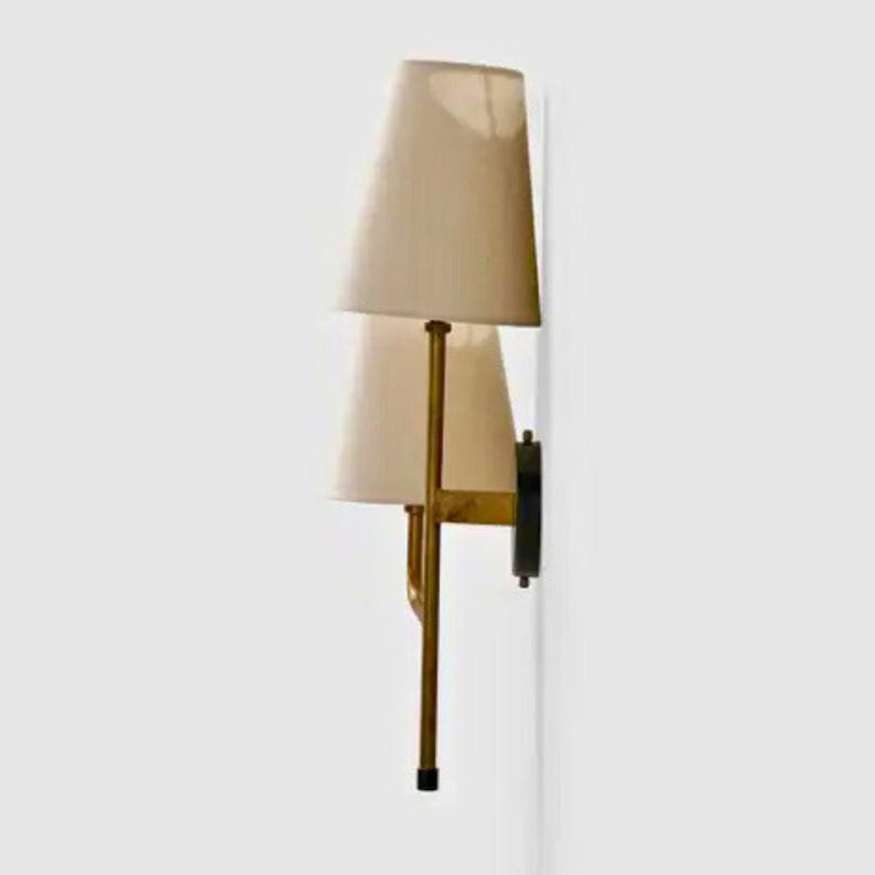 May include: A wall-mounted sconce with a cream-colored lampshade and a brass-colored frame. The sconce has a black mounting plate and a sleek, modern design. The lampshade is cone-shaped and provides soft lighting.
