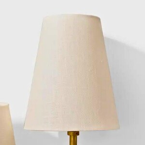 May include: A cream-colored lampshade with a tapered design. The lampshade is made of a textured fabric and is mounted on a gold-colored base. The lamp is designed for indoor use and provides soft, ambient lighting.