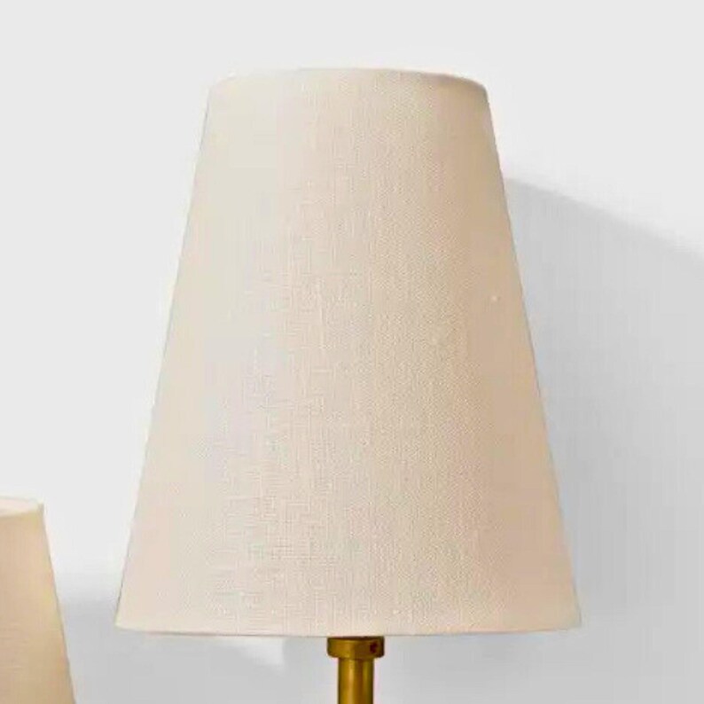 May include: A cream-colored lampshade with a tapered design. The lampshade is made of a textured fabric and is mounted on a gold-colored base. The lamp is designed for indoor use and provides soft, ambient lighting.