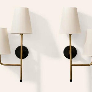 May include: Two wall sconces with off-white tapered shades. Each sconce has a brass-colored arm extending from a black circular base. The sconces are designed with two light fixtures each, creating a symmetrical and elegant lighting arrangement.
