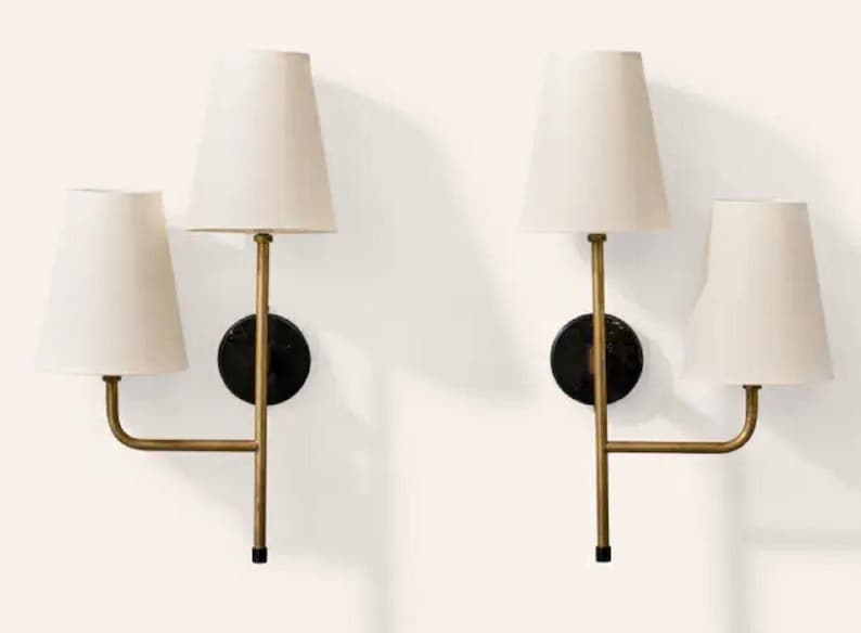 May include: Two wall sconces with off-white tapered shades. Each sconce has a brass-colored arm extending from a black circular base. The sconces are designed with two light fixtures each, creating a symmetrical and elegant lighting arrangement.