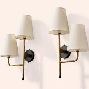 May include: Two wall sconces with off-white tapered shades. The sconces have a gold-toned metal frame with a black circular base. Each sconce has two light fixtures. The lamps are mounted on a white wall.