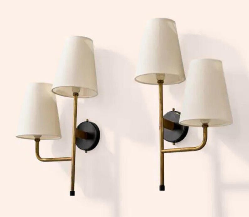 May include: Two wall sconces with off-white tapered shades. The sconces have a gold-toned metal frame with a black circular base. Each sconce has two light fixtures. The lamps are mounted on a white wall.