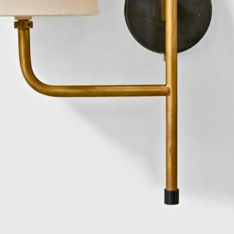 May include: A wall-mounted sconce with a gold-colored metal frame. The design features a curved arm supporting a beige lampshade. A black circular element is attached to the wall, and the base has a black cap.