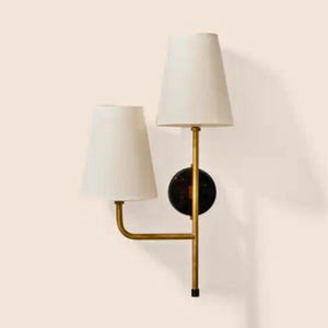 May include: A wall-mounted light fixture with two white lampshades. The fixture has a gold-colored frame and a black circular base. The lampshades are cone-shaped and the fixture is against a light-colored wall.