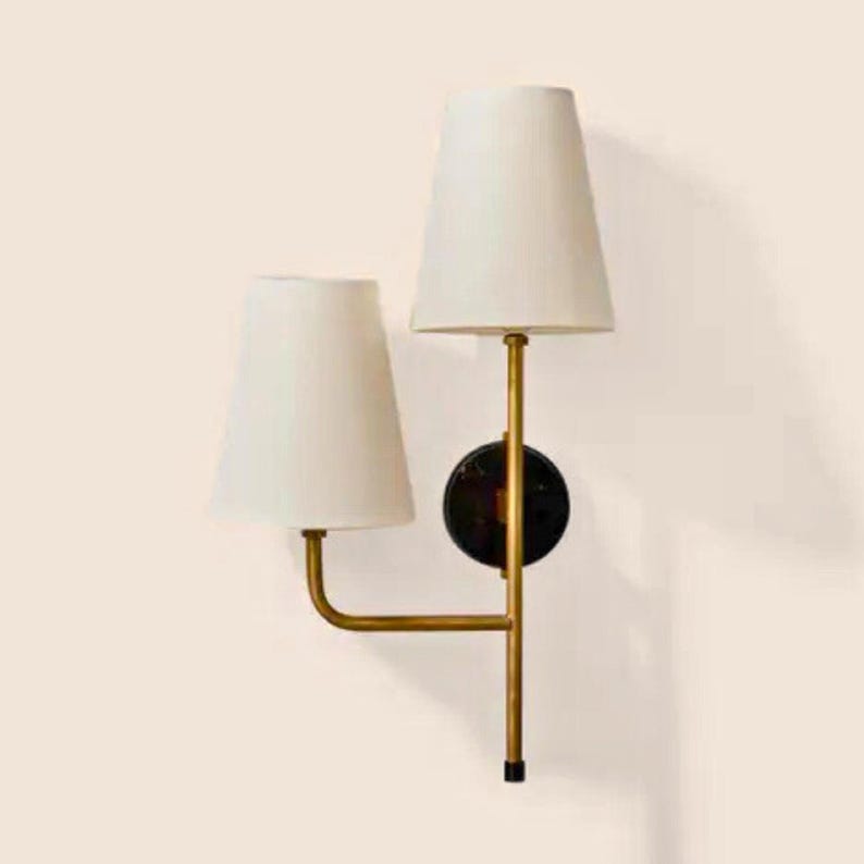 May include: A wall-mounted light fixture with two white lampshades. The fixture has a gold-colored frame and a black circular base. The lampshades are cone-shaped and the fixture is against a light-colored wall.