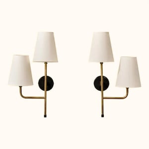 May include: Two wall sconces with white tapered shades and brass-colored arms. Each sconce has a black circular base. The lamps are designed to be mounted on a wall and provide ambient lighting.