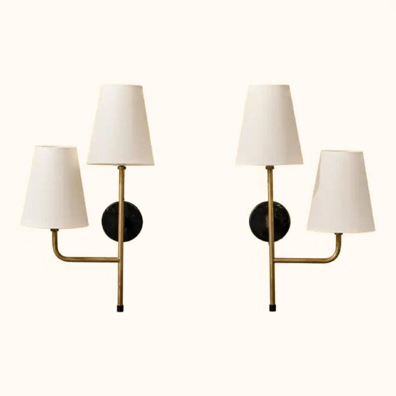 May include: Two wall sconces with white tapered shades and brass-colored arms. Each sconce has a black circular base. The lamps are designed to be mounted on a wall and provide ambient lighting.