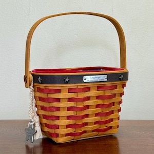 Puede incluir: Una cesta tejida con asa de madera y un patrón rojo y beige. La cesta tiene una banda negra con detalles de estrellas y una etiqueta metálica que dice "Longaberger 2000 Inaugural Basket". Un dije en forma de estrella cuelga de un lado.