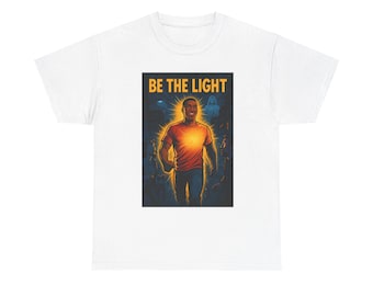 Könnte beinhalten: Weißes T-Shirt mit einer Grafik eines Mannes, der Licht ausstrahlt, und dem goldenen Text "BE THE LIGHT". Der Mann trägt ein orangefarbenes Hemd und blaue Jeans, vor einem dunklen Hintergrund mit einer Menschenmenge.