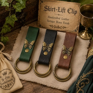 May include: Three skirt-lift clips in green, black, and brown leather, each featuring an antique brass butterfly emblem and a matching brass ring. A rectangular card displays the text Skirt-Lift Clip, Handcrafted Leather, Antique Brass Finish.