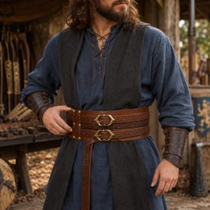 May include: A dark blue long-sleeved tunic worn under a black sleeveless vest, featuring a wide brown leather belt with two brass buckles and matching brown leather bracers on the forearms, all decorated with intricate embossed patterns.