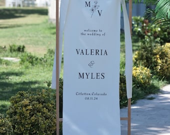 Könnte beinhalten: Ein weißes Hochzeits-Willkommensschild auf einem Holzrahmen. Das Schild zeigt die Initialen "M" und "V" mit floralen Akzenten sowie die Namen "Valeria & Myles" mit Hochzeitsdatum und -ort. Weiße Bänder sind oben gebunden.