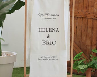 May include: A wooden wedding sign with a white fabric banner. The banner reads "Willkommen" and the names "Helena & Eric" with the date "12. August 2025". The sign is decorated with white bows.
