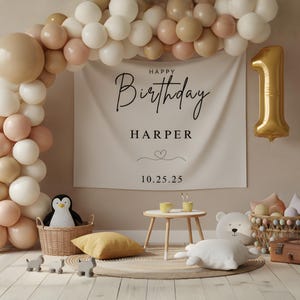 May include: A birthday party scene with a banner that reads "Happy Birthday Harper" and the date 10.25.25. A gold number one balloon hangs to the right. Balloons in white, tan, and pink create an arch. A penguin plush sits in a basket.