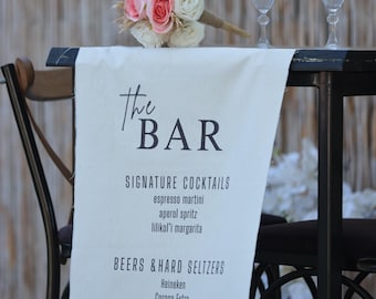 May include: A bar setup with a white linen table runner featuring the text "The BAR" and a list of signature cocktails, beers, and wines. A bouquet of pink and white roses sits atop the table, along with two wine glasses and a decorative candle holder.