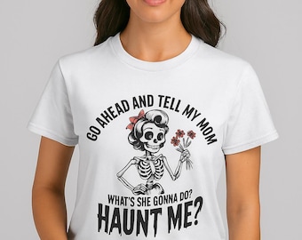 May include: White t-shirt featuring a skeleton graphic with a red bow and flowers. The text reads "GO AHEAD AND TELL MY MOM WHAT'S SHE GONNA DO? HAUNT ME?" A fun, spooky design.