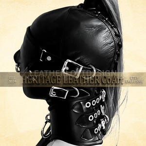 Op de afbeelding: Een zwarte lederen hoofdbedekking met een opening voor een paardenstaart, ge&iuml;ntegreerd oogmasker en een brede halsband. Het ontwerp bevat meerdere zilverkleurige metalen gespen, D-ringen en vetersluitingen aan de zijkant. Tekst Heritage Leather CoArt.