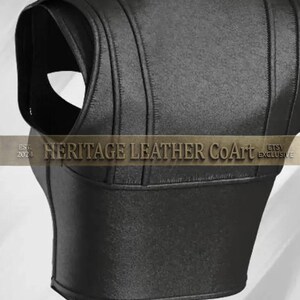 Puede incluir: Un chaleco negro sin mangas con paneles cosidos verticales y un dobladillo en punta. La prenda muestra el texto "EST. 2024 HERITAGE LEATHER CoArt ETSY EXCLUSIVE" en la parte central. El dise&ntilde;o incluye aberturas anchas para los brazos y un corte estructurado.