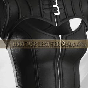 Puede incluir: Un top negro sin mangas con cuello alto, cierre de hebilla plateada, abertura en el pecho y cremallera frontal completa. La prenda muestra el texto HERITAGE LEATHER COArt EST 2024 ETSY EXCLUSIVE en la parte central del dise&ntilde;o.