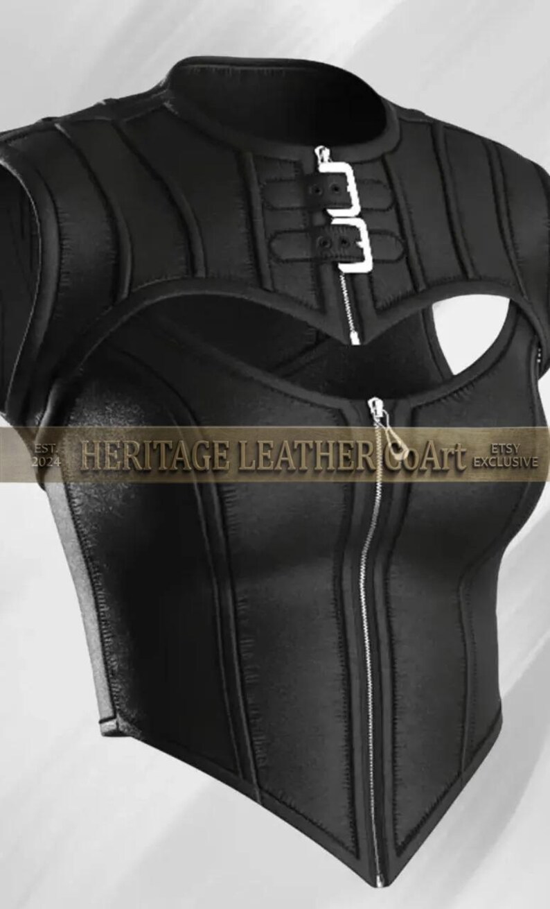Puede incluir: Un top negro sin mangas con cuello alto, cierre de hebilla plateada, abertura en el pecho y cremallera frontal completa. La prenda muestra el texto HERITAGE LEATHER COArt EST 2024 ETSY EXCLUSIVE en la parte central del dise&ntilde;o.