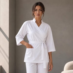 May include: A solid white scrub set consisting of a wrap-style tunic with three-quarter length sleeves, a tie waist, and a front pocket, paired with matching straight-leg trousers and white sneakers, creating a clean and uniform professional look.