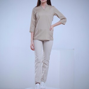 May include: A beige uniform set, including a top with three-quarter sleeves and a V-neck, paired with matching pants. The outfit is worn with white sneakers. The model is standing on a white cube against a light background.